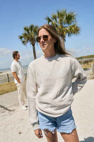A woman in The Stamp Crewneck Sweatshirt, sunglasses, and denim shorts stands outside near palm trees, with a man in white in the background under a blue sky.