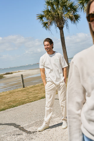A man in The Stamp Tee, a relaxed fit retro unisex t-shirt, and light pants stands smiling on a sunny path near the beach with a palm tree and water behind him. Part of another person is seen in the right foreground.