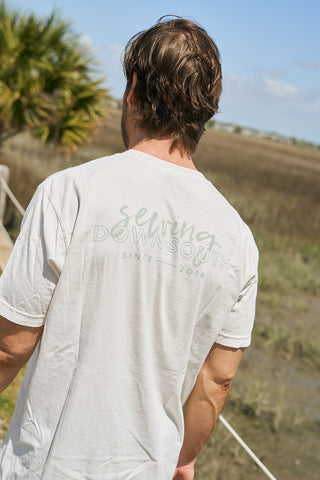 A man with light brown hair stands outdoors, facing away from the camera, wearing The Stamp Tee—a cream-colored retro unisex shirt that reads "serving DOWN SOUTH SINCE 2018." Marshland and a palm tree appear in the background.