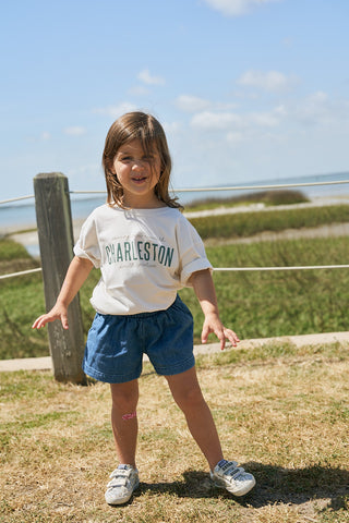 A smiling toddler stands on the grass by the beach, wearing a Youth Charleston Tee, blue shorts, sneakers, and a pink bandage on their leg, with the ocean and blue sky in the background.