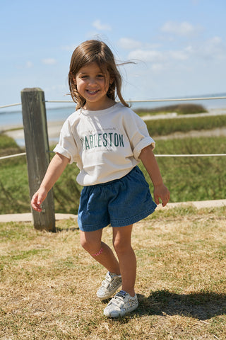 A young toddler smiles outdoors in the sun, wearing a white Youth Charleston Tee with a boxy fit, blue skirt, white sneakers, and a pink knee bandage. Grass, a wooden post, fence, and water appear in the background.