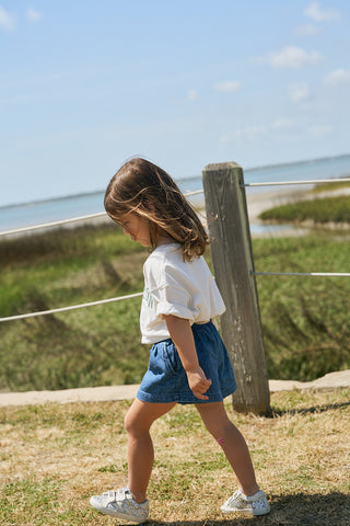 A toddler with shoulder-length brown hair walks outdoors on a sunny day, wearing a white Youth Charleston Tee, blue shorts, and sneakers. Grass, water, and a wooden post with rope are visible in the background.
