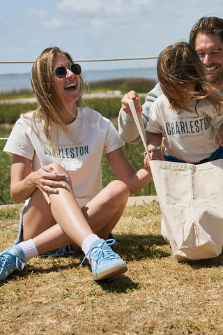 A woman, man, and child sit outdoors on dry grass under a sunny sky, all smiling and wearing matching The Charleston Women's Tee shirts with ribbed collars. The child looks into a large white tote bag amid greenery.