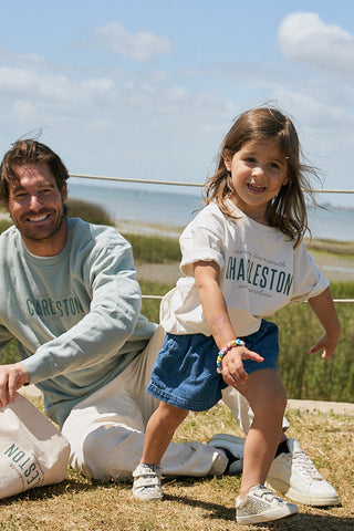 A smiling man in a light green sweatshirt poses by the water with a young girl wearing blue shorts and a white Youth Charleston Tee.