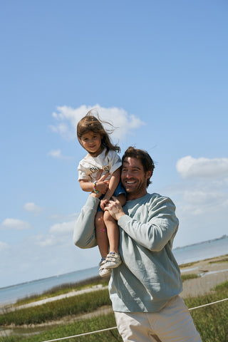 A man lifts a toddler onto his shoulders near a beach. The girl, wearing the Youth Charleston Tee, looks at the camera as her hair blows in the wind. Blue sky and grassy shoreline are visible in the background.