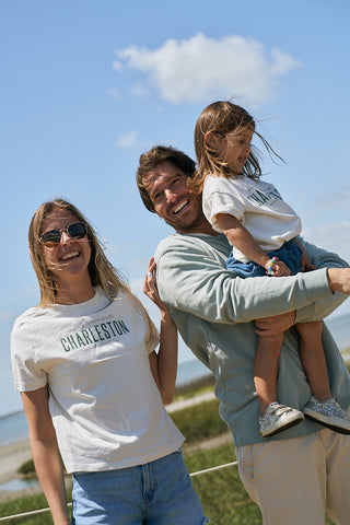 A smiling family of three stands outdoors on a sunny day. The woman and young girl wear The Charleston Women's Tee with a ribbed collar, as the man holds the girl. All appear happy with blue sky and water in the background.