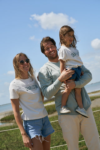 A smiling man and woman stand outdoors with a young girl, all in casual wear. The woman is wearing The Charleston Women's Tee, with water and greenery in the background on a sunny day.