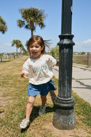 A young child with brown hair, wearing the Youth Charleston Tee and blue shorts, joyfully runs around a lamppost in a sunny outdoor park with palm trees and a clear sky.