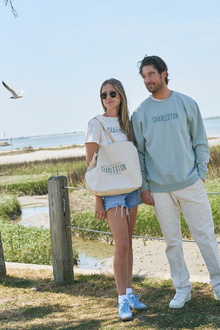 A woman with long hair and sunglasses stands outdoors near a beach in a casual Charleston sweatshirt, carrying The Charleston Tote with canvas webbed handles. A man stands beside her as a seagull soars overhead.