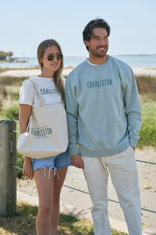 A woman and man stand smiling near the beach, holding hands in The Charleston Crewneck Sweatshirts. She wears sunglasses, denim shorts, carries a tote and crossbody; he pairs his sweatshirt with white pants.