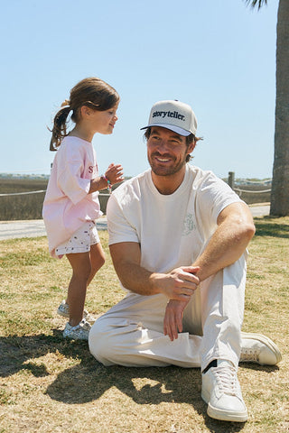 A man in white sits smiling on the grass as a young girl outdoors in a sunny setting stands beside him, wearing the Youth Storyteller Tee, a pink toddler t-shirt made of soft combed ring-spun cotton.