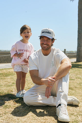 A smiling man kneels on grass in a boxy cotton tee beside a cheerful girl in a soft combed ring-spun cotton Youth Storyteller Tee. Both wear white storyteller shirts, enjoying the outdoors together.