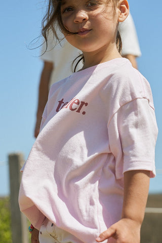 A young child with long brown hair wears the boxy fit Youth Storyteller Tee with "trier." in red text, standing outdoors on a sunny day with a blurred person and blue sky behind them.