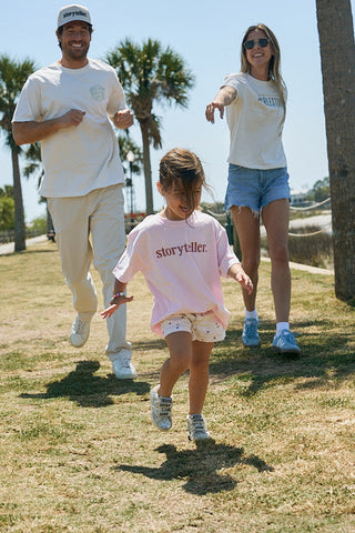 A young child wearing the Youth Storyteller Tee runs on grass as two adults chase playfully behind, outdoors under sunny skies with palm trees in the background.