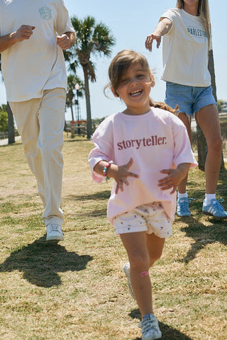 A young girl smiles while running on grass in a pink Youth Storyteller Tee. Two adults, partially visible, follow behind her on a sunny day with palm trees in the background.