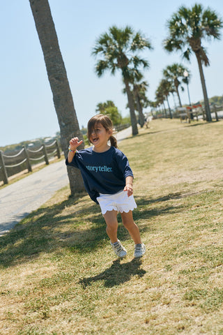 A young girl in a navy Youth Storyteller Tee and white shorts runs happily along a grassy path bordered by palm trees on a sunny day.