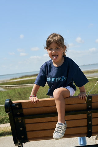 A young girl playfully sits on a bench by the water, smiling under a sunny blue sky. She wears the Youth Storyteller Tee in navy, paired with white shorts and sneakers for a carefree summer look.