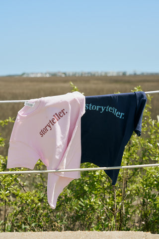 Two Youth Storyteller Tees—one light pink, one navy blue, both boxy fit cotton with “storyteller.” on the front—hang side by side over a white rope outdoors, surrounded by greenery and open fields.
