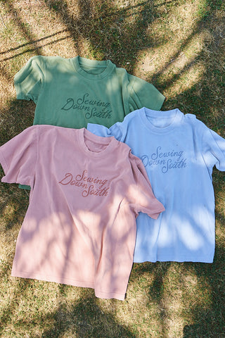 Three Rainbow Row Script Tees in green, pink, and light blue with "Sewing Down South" on the front are laid out on sunlit grass. They feature an oversized boxy fit, heavyweight jersey fabric, and tree branch shadows across them.