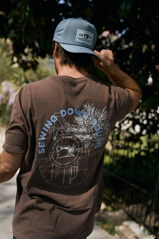 A person wearing a brown T-shirt with a circular white and blue graphic and The Silicone Patch Hat in Breaker Blue stands outdoors, facing away from the camera, surrounded by greenery and sunlight.
