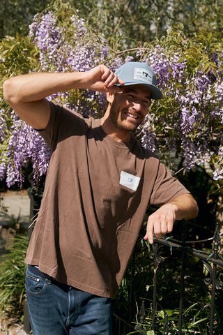 A smiling man in a brown t-shirt tips The Silicone Patch Hat in Breaker Blue while standing outdoors, leaning on a black metal fence before blooming wisteria vines with vibrant purple flowers.