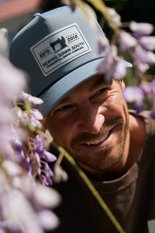 A smiling man wearing The Silicone Patch Hat in Breaker Blue is partially obscured by purple flowers in sunlight.