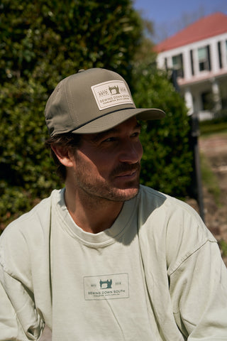 A man wearing an olive green Silicone Patch Hat and a light green Sewing Down South shirt sits outdoors, surrounded by greenery and a white house in the blurred background.