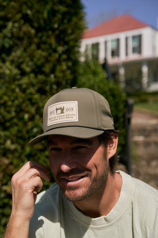 A man with light facial hair, wearing an olive green The Silicone Patch Hat, smiles outdoors in front of greenery and a white house, dressed in a light shirt.