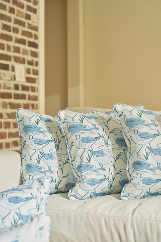 Three Sandpiper Pillows, featuring blue shorebird and plant designs, rest on a light sofa. A brick wall and a beige painted wall in the background complete the coastal decor vibe.