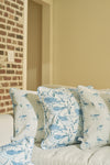 Three Sandpiper Pillows with blue bird and floral patterns rest on a light sofa, while brick and beige walls in the background enhance the charm of this seaside-inspired decor.