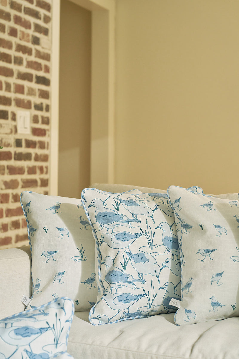 Three Sandpiper Pillows with blue bird and floral patterns rest on a light sofa, while brick and beige walls in the background enhance the charm of this seaside-inspired decor.