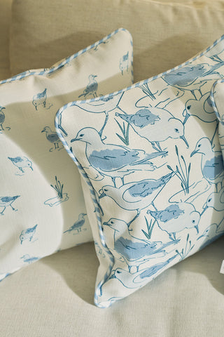 Two cream-colored Sandpiper Pillows with blue shorebird and grass designs rest on a beige couch. One pillow shows larger birds, the other a smaller repeating pattern—ideal for adding seaside charm to your decor.