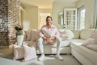 A man in a light pink shirt and white pants sits smiling on a white sofa, holding the Pink Lattice Pillow, surrounded by blush-toned pillows in a bright, cozy living room with brick and white walls.