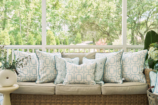 A wicker sofa with beige cushions and ocean-inspired style features white and light blue patterned pillows, including the Starfish Trio with Beach Lattice Lumbar Pillow. It sits on a porch with a white railing, surrounded by lush greenery.