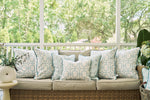 A wicker sofa with beige cushions and ocean-inspired style features white and light blue patterned pillows, including the Starfish Trio with Beach Lattice Lumbar Pillow. It sits on a porch with a white railing, surrounded by lush greenery.
