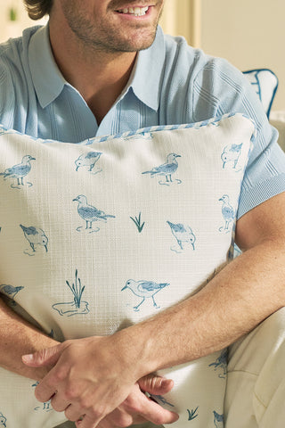 A man in a light blue shirt sits and hugs The Sandpiper Pillow, featuring blue sandpipers and coastal grasses. Only the lower half of his smiling face is visible.