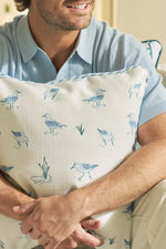 A man in a light blue shirt sits and hugs The Sandpiper Pillow, featuring blue sandpipers and coastal grasses. Only the lower half of his smiling face is visible.