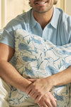 A person in a light blue shirt smiles while hugging The Sandpiper Pillow, featuring blue and white sandpiper and grass illustrations. Only their lower face shows, adding charm to this coastal decor scene.