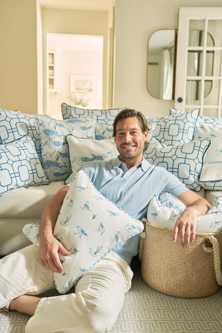 A smiling man in a light blue shirt and beige pants sits on a couch in a cozy, well-lit living room, holding The Sandpiper Pillow and resting his arm on a woven basket amid blue and white coastal pillows.