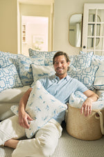 A smiling man in a light blue shirt and beige pants sits on a couch in a cozy, well-lit living room, holding The Sandpiper Pillow and resting his arm on a woven basket amid blue and white coastal pillows.
