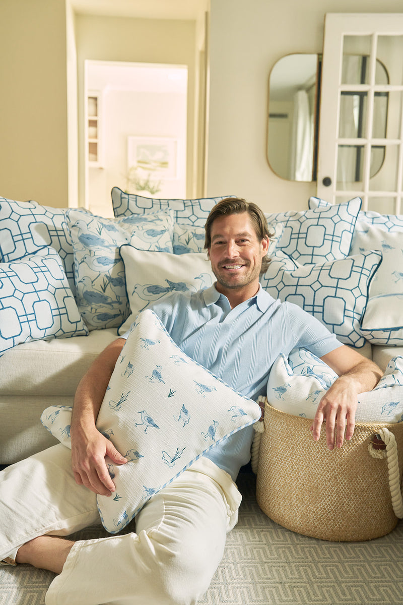A smiling man in a light blue shirt and beige pants sits on a couch in a cozy, well-lit living room, holding The Sandpiper Pillow and resting his arm on a woven basket amid blue and white coastal pillows.