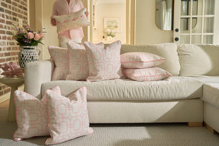 A cream-colored sectional sofa is adorned with pink and white patterned pillows, including the Pink Lattice Pillow in blush. In the cozy living room background, a person stands holding a pillow featuring pink flamingos.