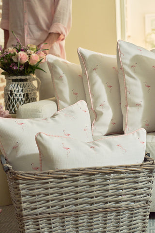 A wicker basket holds the Flamingo Scatter Oversized Pillow with pink patterns beside a cream sofa, while a vase of flowers and someone in a pale pink shirt appear in the softly lit background.