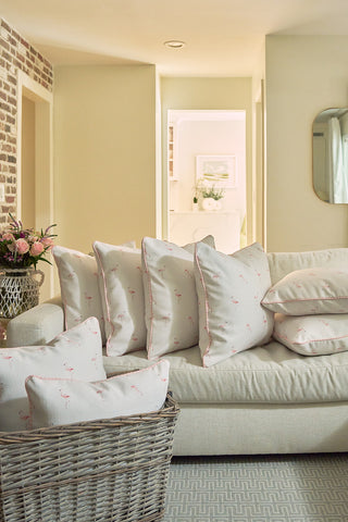 A cozy living room features a cream sofa with white pillows accented in pink, including the Flamingo Scatter Oversized Pillow. More pillows are stored in a wicker basket, and a bouquet of flowers sits in warm, natural light.