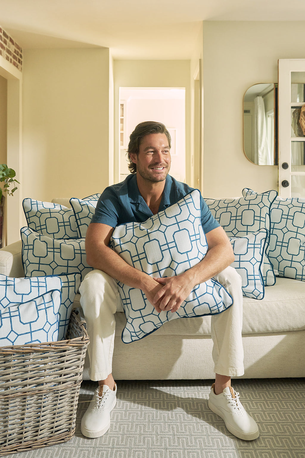 A man sits on a beige sofa, smiling and holding the Blue Lattice Oversized Pillow. Several large pillows and a basket of pillows are nearby in a bright, modern living room with coastal blue accents.