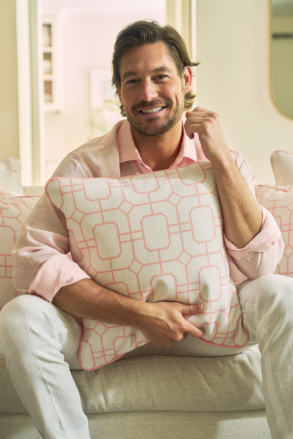 A smiling man with light brown hair and a beard, in a pale pink shirt and white pants, sits on a light sofa holding the Pink Lattice Pillow, which features timeless charm and a classic geometric lattice design.