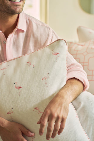 A person in a light pink shirt smiles while holding the Flamingo Scatter Oversized Pillow, featuring small pink flamingos on durable fabric. Another pillow with an elevated twisted welt is seen in the background.