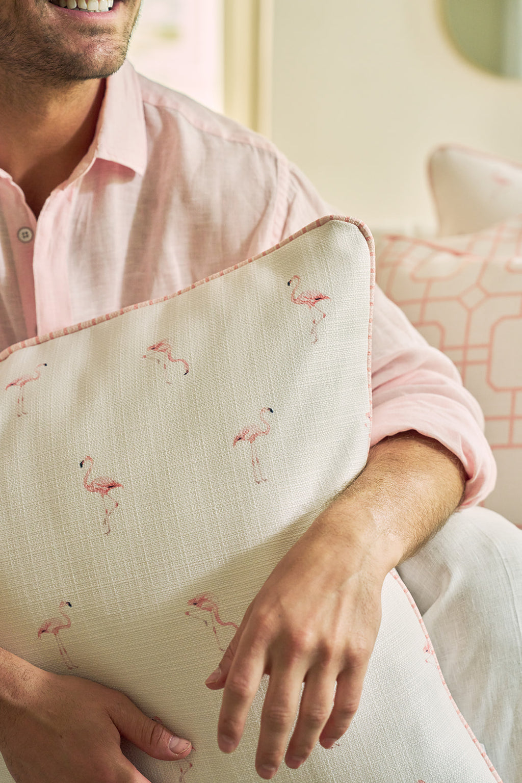 A person in a light pink shirt smiles while holding the Flamingo Scatter Oversized Pillow, featuring small pink flamingos on durable fabric. Another pillow with an elevated twisted welt is seen in the background.