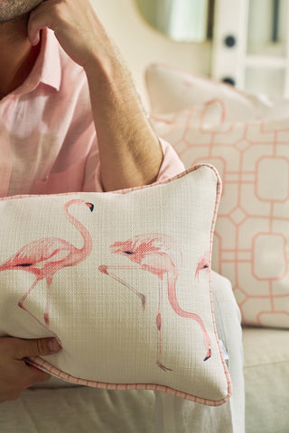 A person in a light pink shirt holds a Flamingo Cluster Extended Lumbar Pillow with pink flamingo illustrations, adding coastal charm, while a soft pink geometric-patterned pillow is visible in the background.