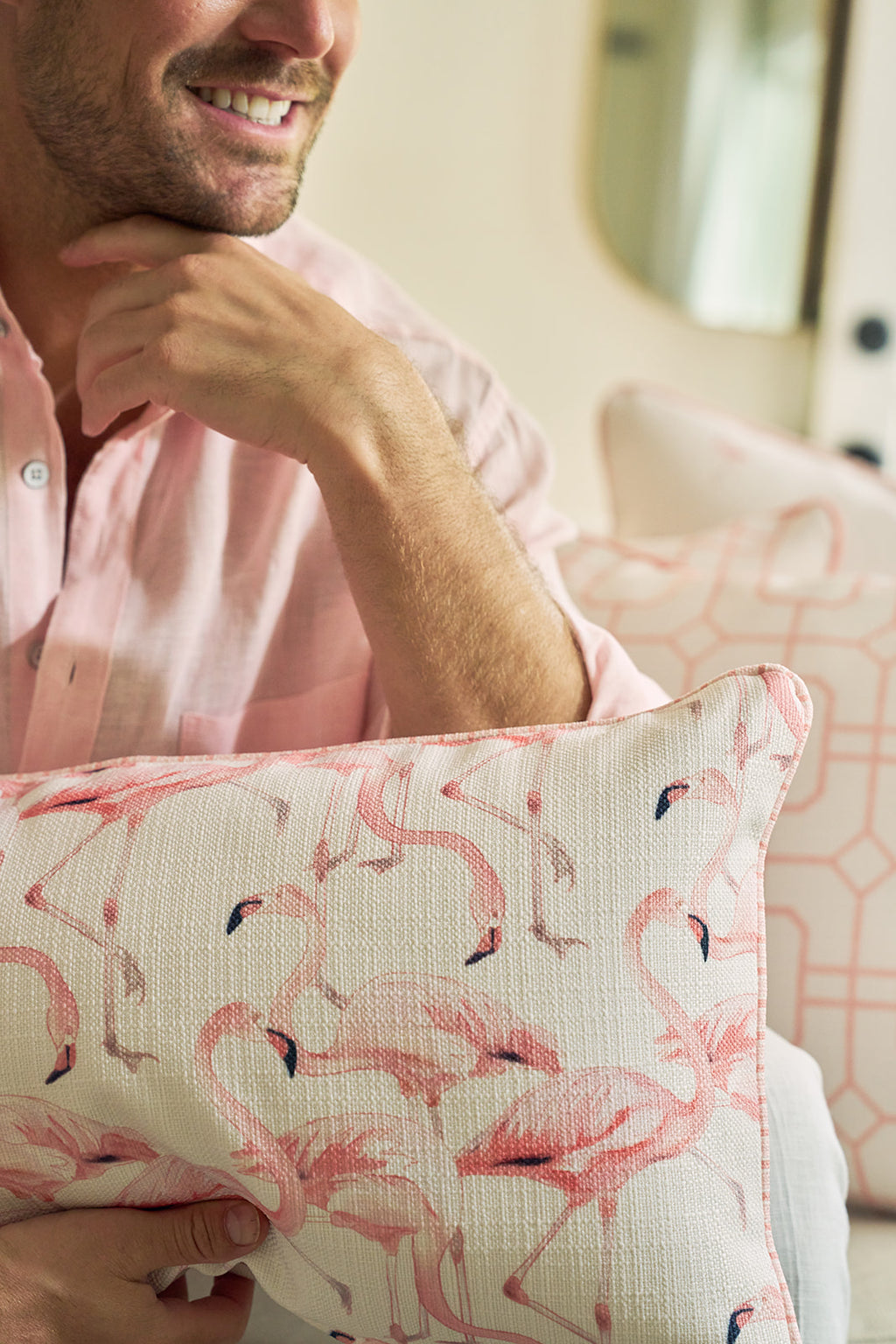A smiling person in a light pink shirt holds the Flamingo Cluster Extended Lumbar Pillow, featuring the iconic Sewing Down South® pattern. White and pink geometric pillows in the background add to the coastal charm.